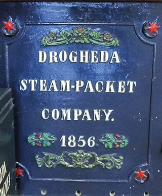No alto: Uma placa decorativa de metal da empresa de transporte marítimo Steam-Packet de Drogheda, fundada em 1826 e que até meados do século foi a empresa marítima dominante da cidade, é ornamentada em cada um dos ângulos com a insígnia de crescente e estrela da cidade (que aqui mostra uma estrela de seis pontas).