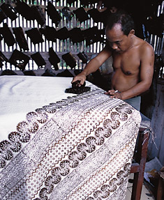An artisan applies a hot wax pattern using a hand block at Apip’s Batik in Jogjakarta; other blocks hang on the louvered wall. 