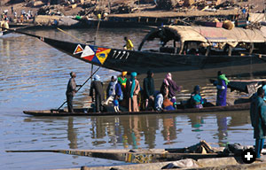 His passengers carefully balancing, a boatman poles his pirogue up to the Niger's embankment at Mopti. Behind the pirogue, a long-distance cargo and passenger pinasse is docked.