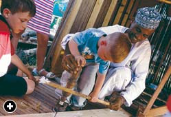An Omani shipwright shows a youngster how to use a traditional bow drill. 