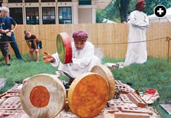 Before a performance, a musician toasts drums around a small fire to tune them, checking the tone from time to time as they warm. 