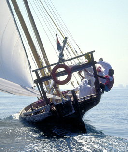 With its wind-filled lateen sail ballooning, a racing dhow heels in a fresh breeze off Dubai. To make sure the boat does not lean too much, which would slow its speed, crew members hike out over the rail. By moving their body weight as far as possible toward the direction of the wind, they aim to counter the force of the wind as it pushes against the sail.