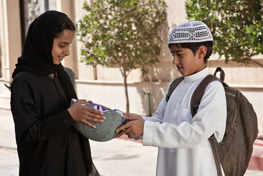 Em seu primeiro gesto daquilo que se tornará um admirável apoio à determinação de Wadjda em aprender a andar de bicicleta, Abdullah lhe dá um capacete. Ao lado, à esquerda: No Festival Internacional de Cinema de Dubai de 2012, Waad Mohammed recebe o prêmio de melhor atriz de longa-metragem das mãos do HH Sheikh Mansoor bin Mohammed bin Rashid Al Maktoum.